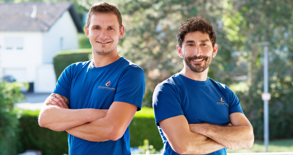 Zwei lächelnde Männer in blauen T-Shirts, stehen mit verschränkten Armen im Freien.